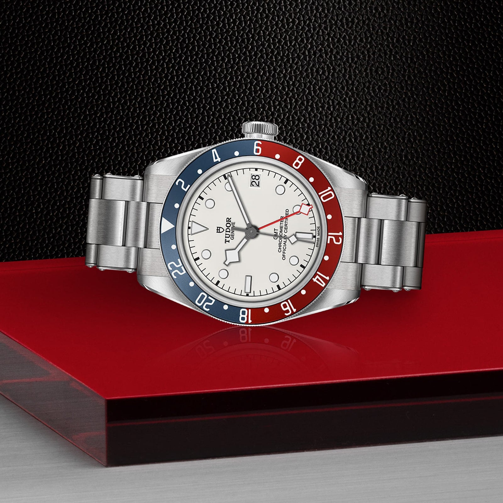 The TUDOR Black Bay GMT, featuring a silver case, red and blue bezel, opaline dial, and stainless steel band, is displayed on a red and black surface with a textured black background—a striking tribute to its iconic design.