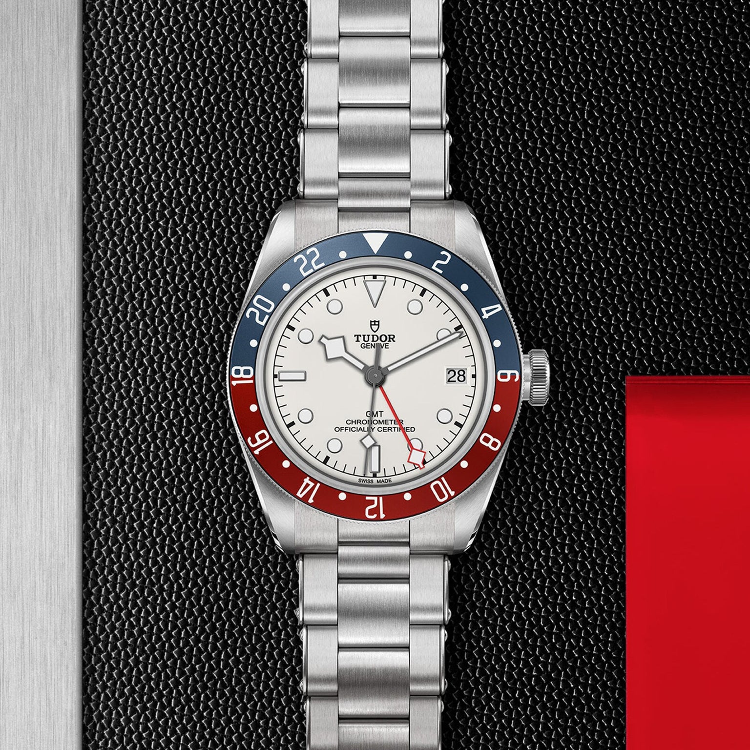 The TUDOR Black Bay GMT wristwatch features a silver metal band and a red and blue bezel, showcased on a textured black surface.