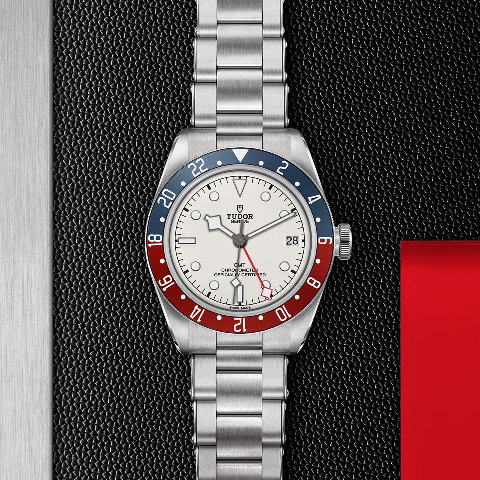 The TUDOR Black Bay GMT wristwatch features a silver metal band and a red and blue bezel, showcased on a textured black surface.