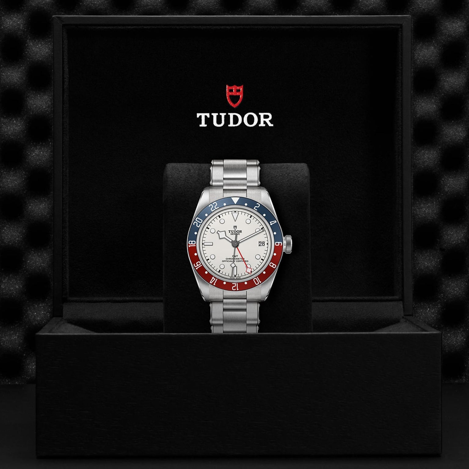 A TUDOR Black Bay GMT wristwatch with a red and blue bezel, opaline dial, and silver band is displayed in an open black TUDOR box, highlighting its iconic GMT function.