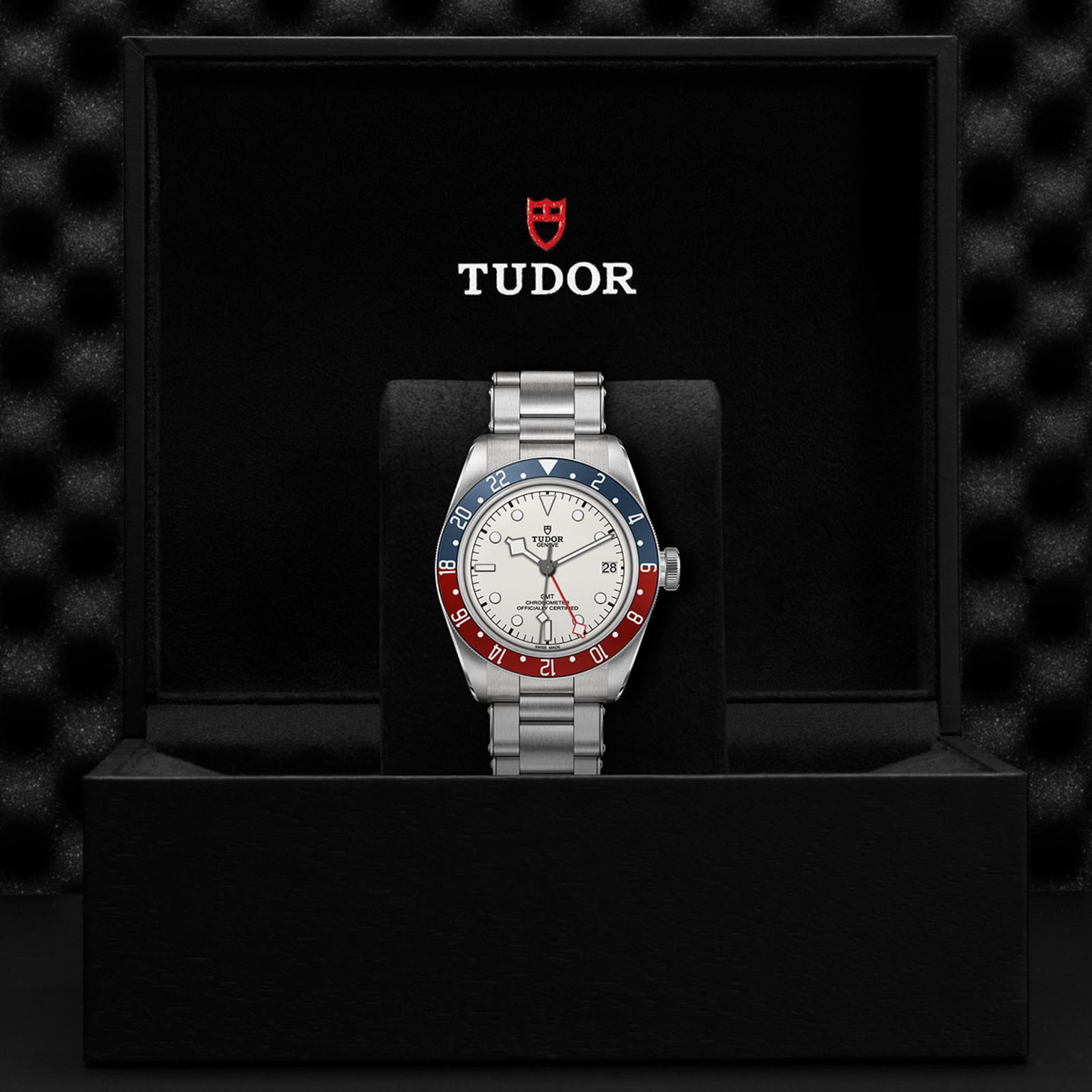 A TUDOR Black Bay GMT wristwatch with a red and blue bezel, opaline dial, and silver band is displayed in an open black TUDOR box, highlighting its iconic GMT function.