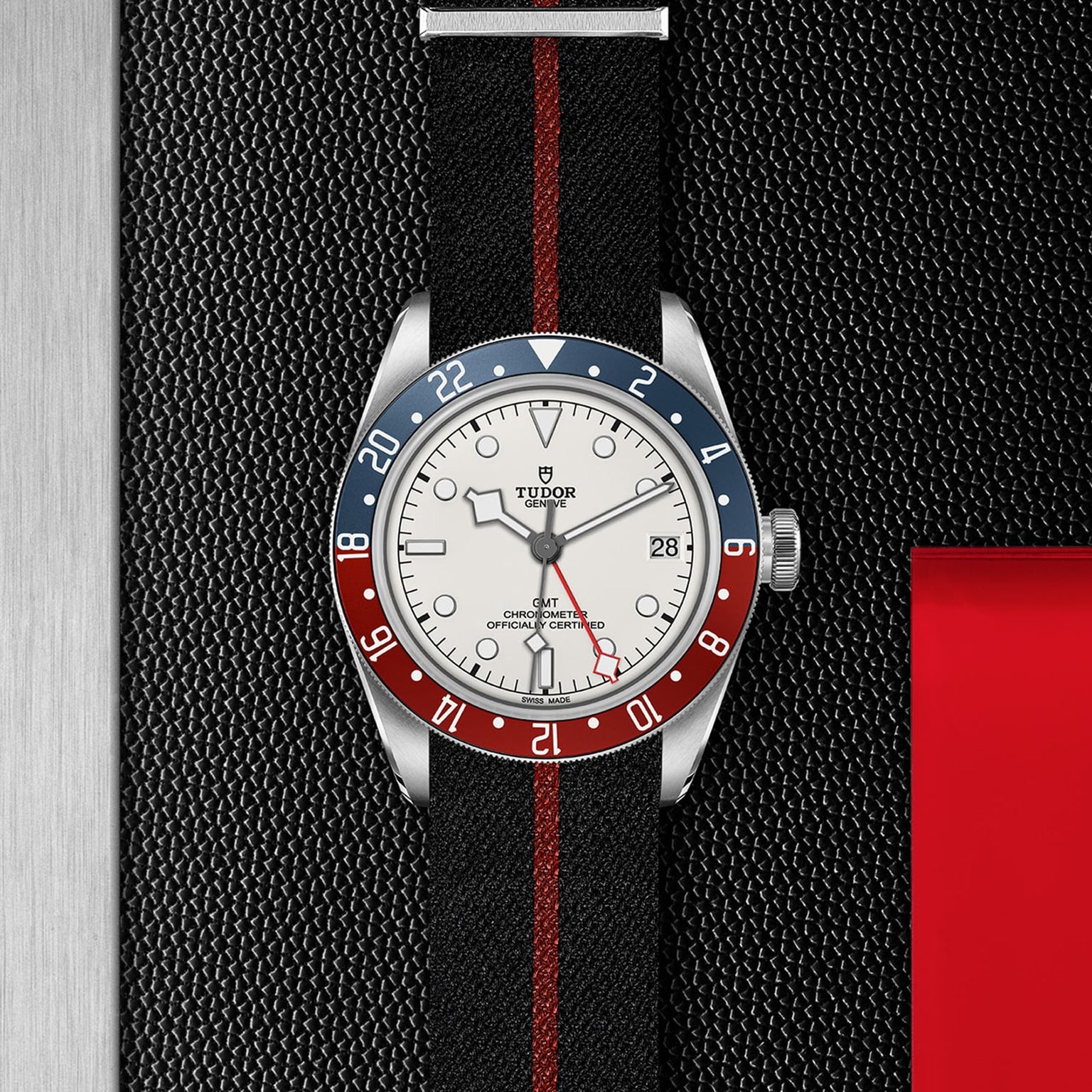 The TUDOR Black Bay GMT wristwatch features a black strap with a red stripe, blue and red bezel, opaline dial, and date display. Shown on a textured black and silver background, it’s ideal for stylishly tracking multiple time zones.