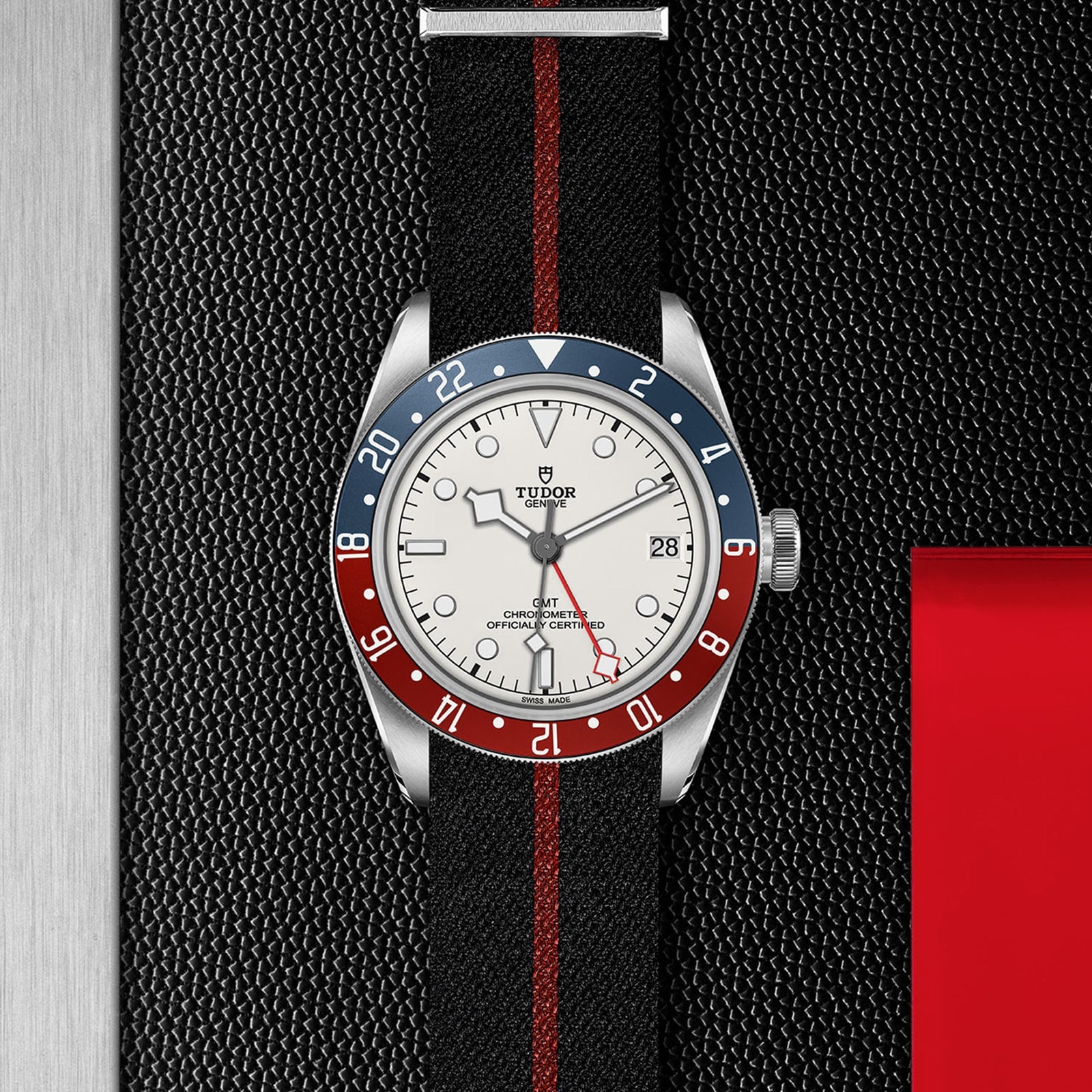 The TUDOR Black Bay GMT wristwatch features a black strap with a red stripe, blue and red bezel, opaline dial, and date display. Shown on a textured black and silver background, it’s ideal for stylishly tracking multiple time zones.