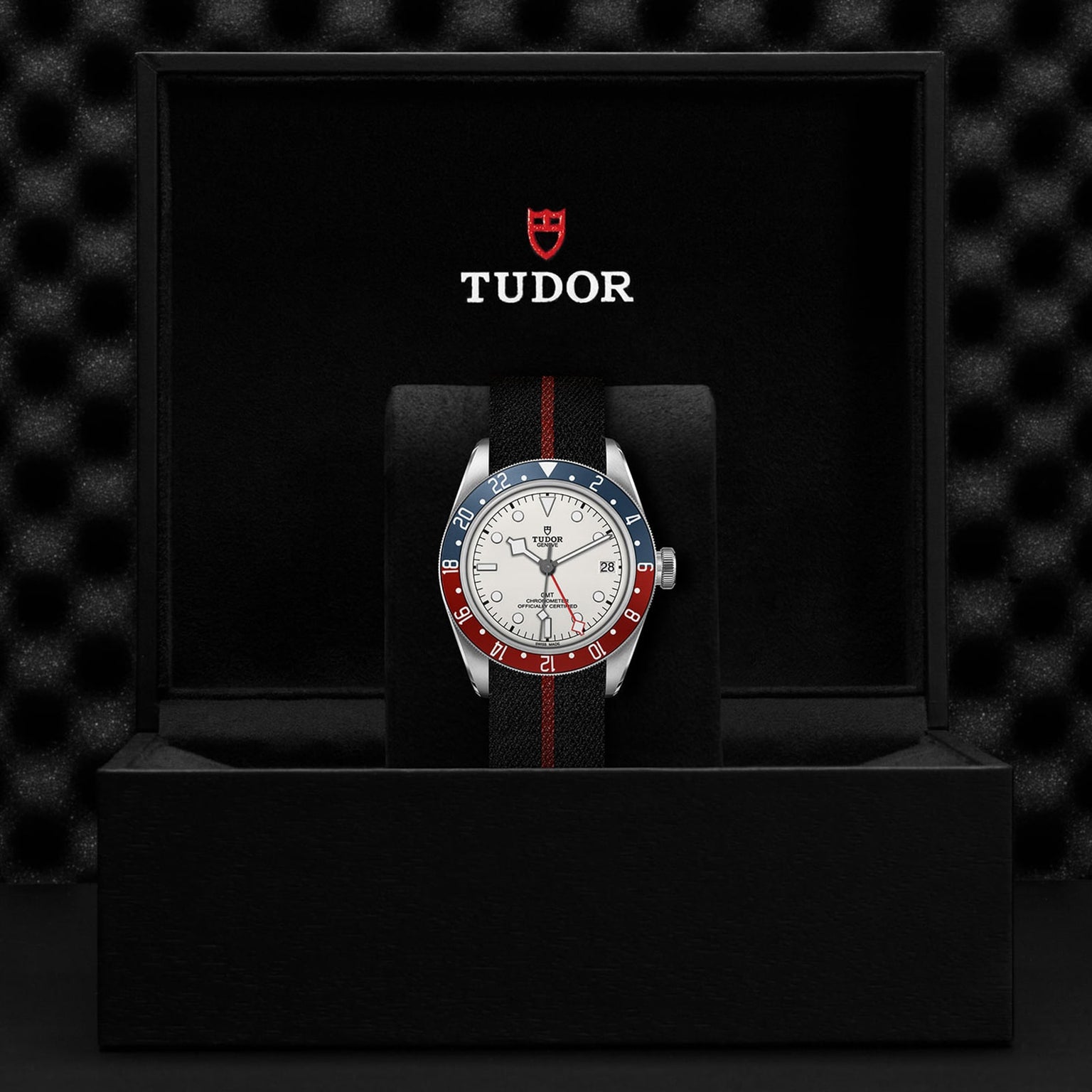 A TUDOR Black Bay GMT wristwatch with a red and blue bezel, black strap, and opaline dial stands upright in a black box bearing the TUDOR logo on the inner lid—ideal for stylishly tracking multiple time zones.