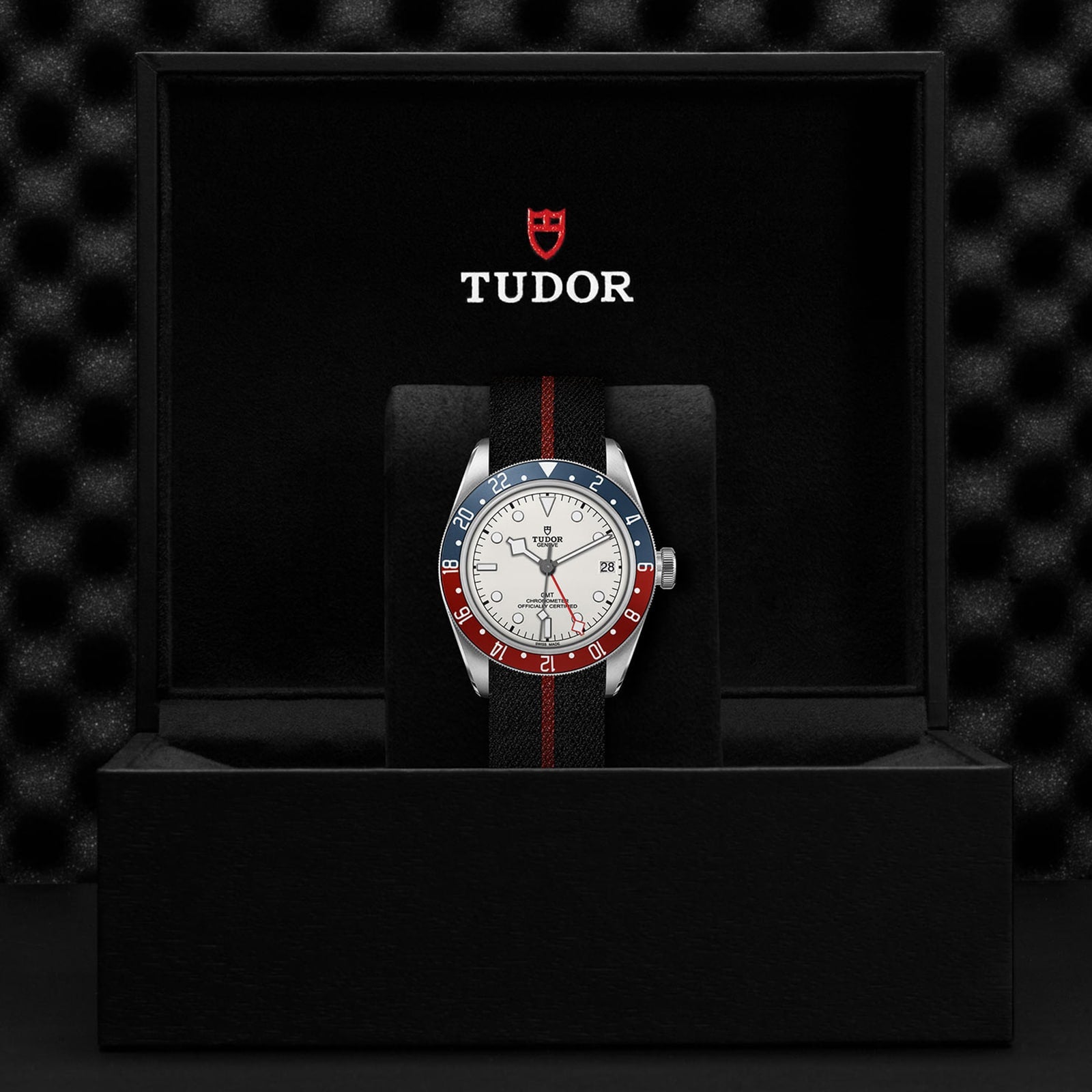 A TUDOR Black Bay GMT wristwatch with a red and blue bezel, black strap, and opaline dial stands upright in a black box bearing the TUDOR logo on the inner lid—ideal for stylishly tracking multiple time zones.
