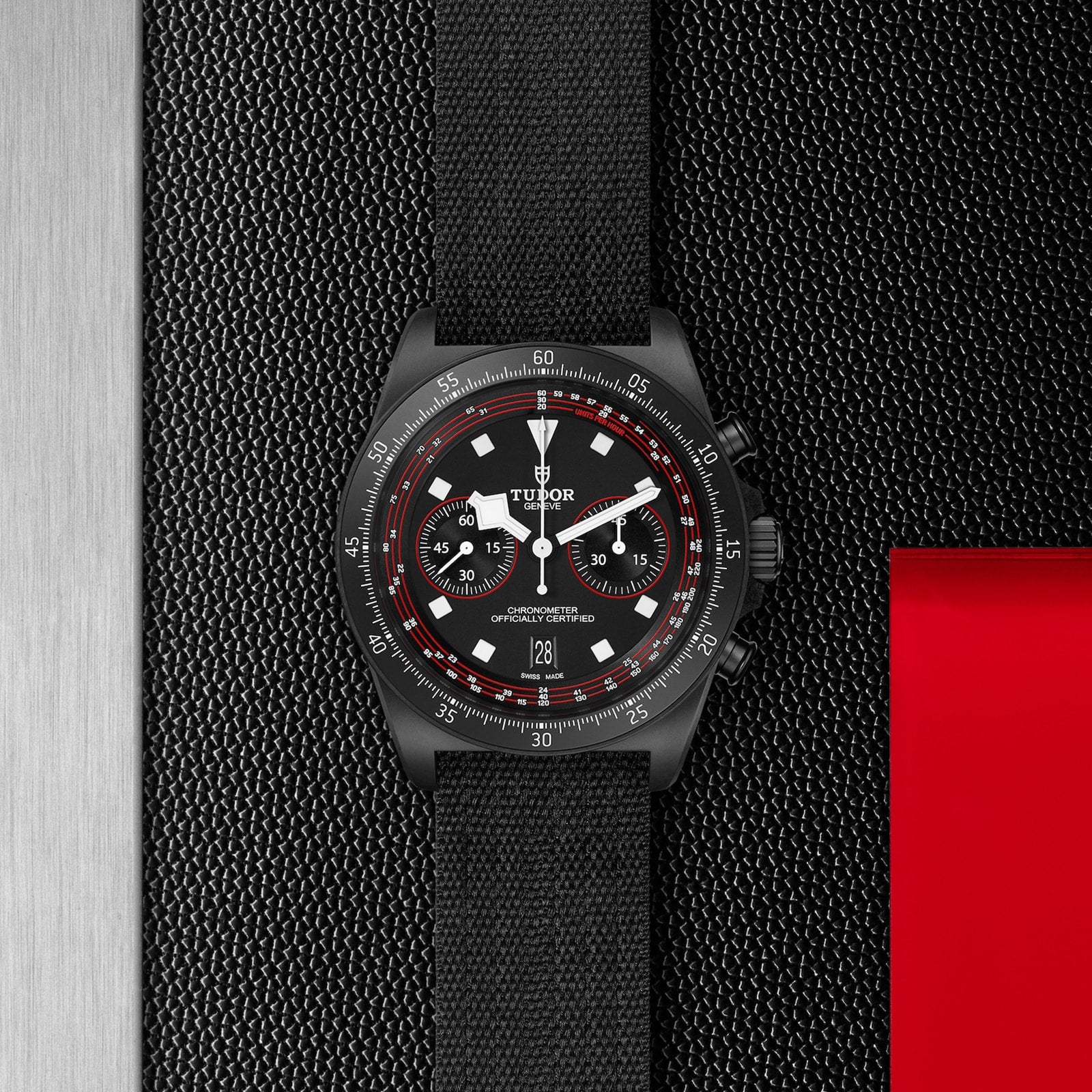 The TUDOR Pelagos FXD Chrono, a black cycling-themed chronograph with red accents and a black strap, is showcased on a textured black background featuring red details.