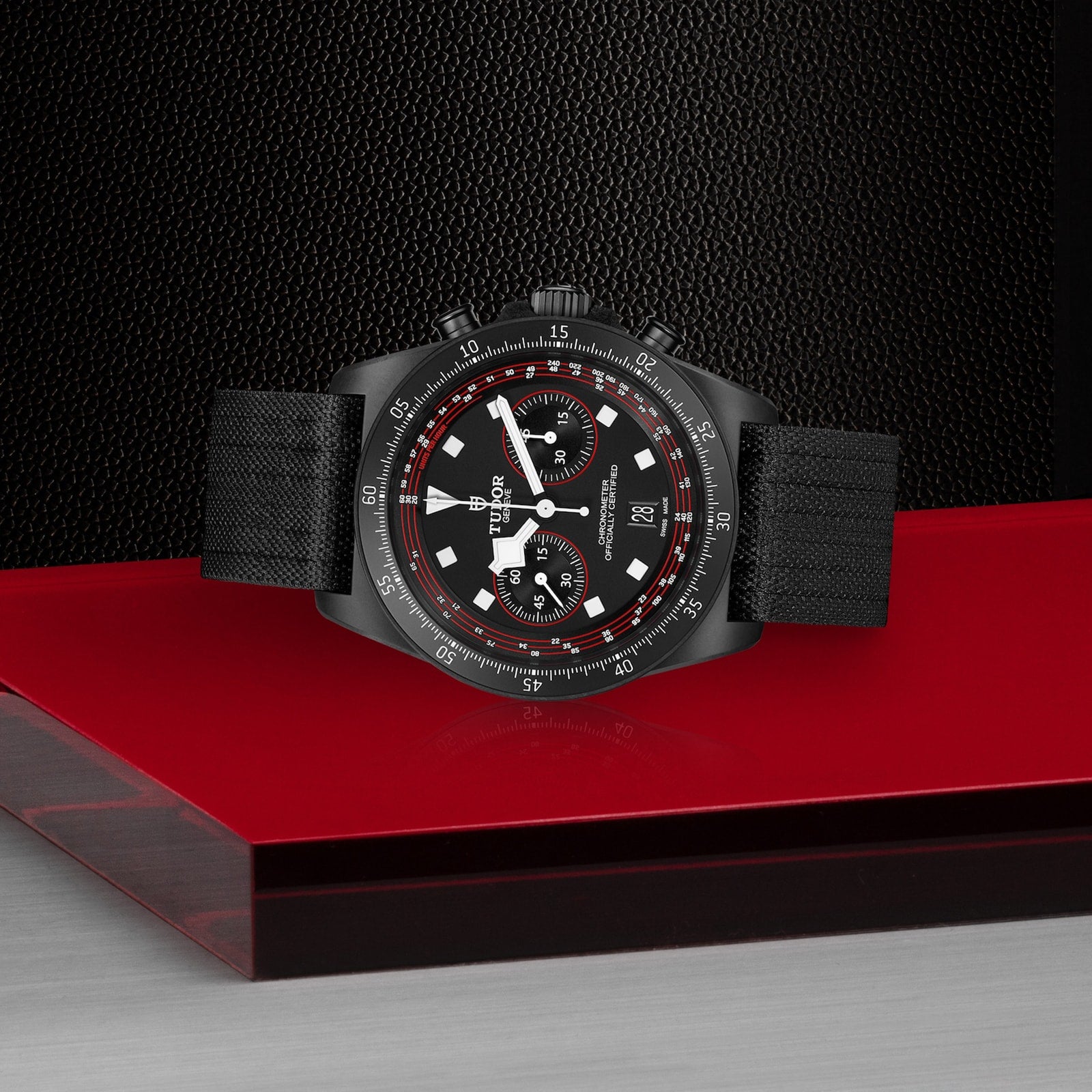 The TUDOR Pelagos FXD Chrono is a black cycling-inspired chronograph watch with red accents and a cycling tachymeter, set on a red and silver surface against a textured black background.