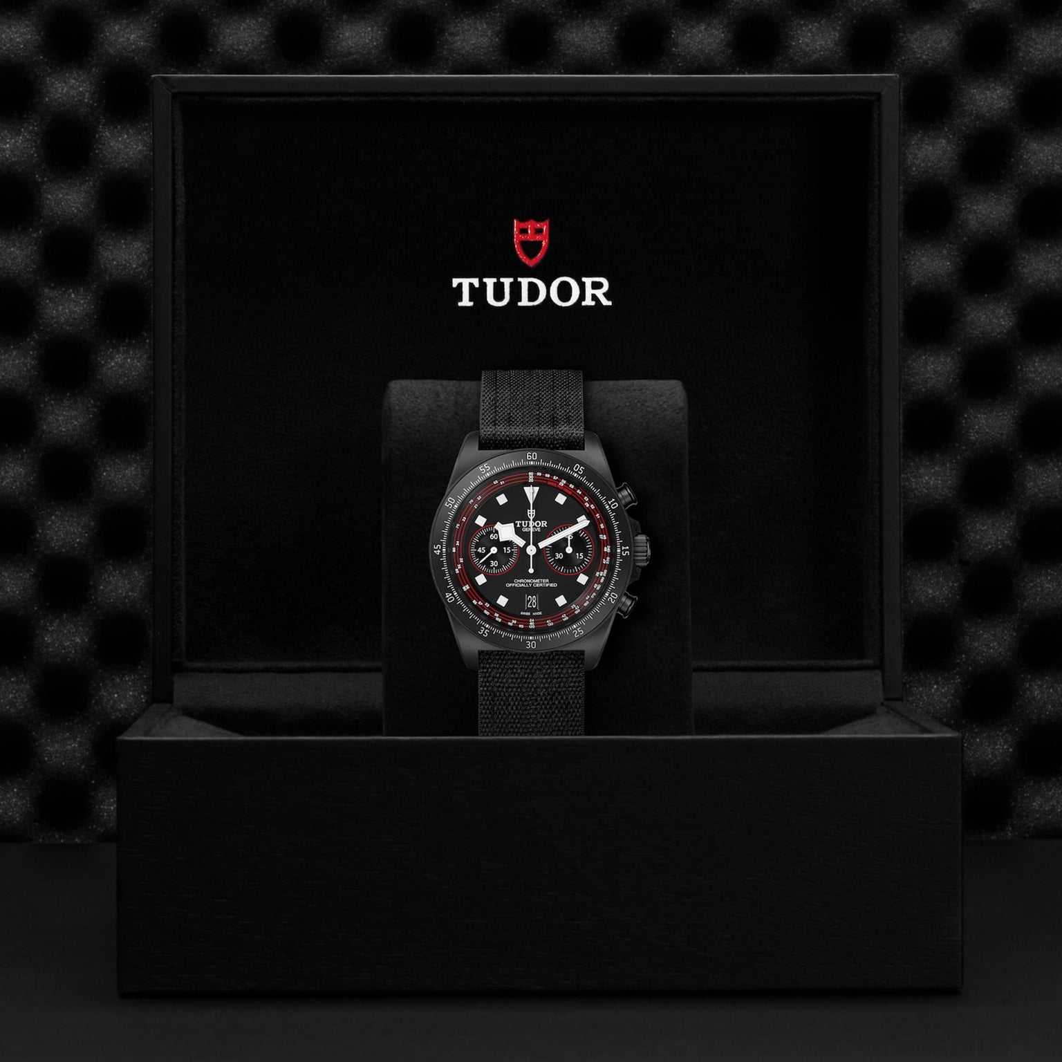 A TUDOR Pelagos FXD Chrono with a black strap and red-accented bezel stands upright in a black TUDOR presentation box, showcasing its cycling-specific tachymeter scale.