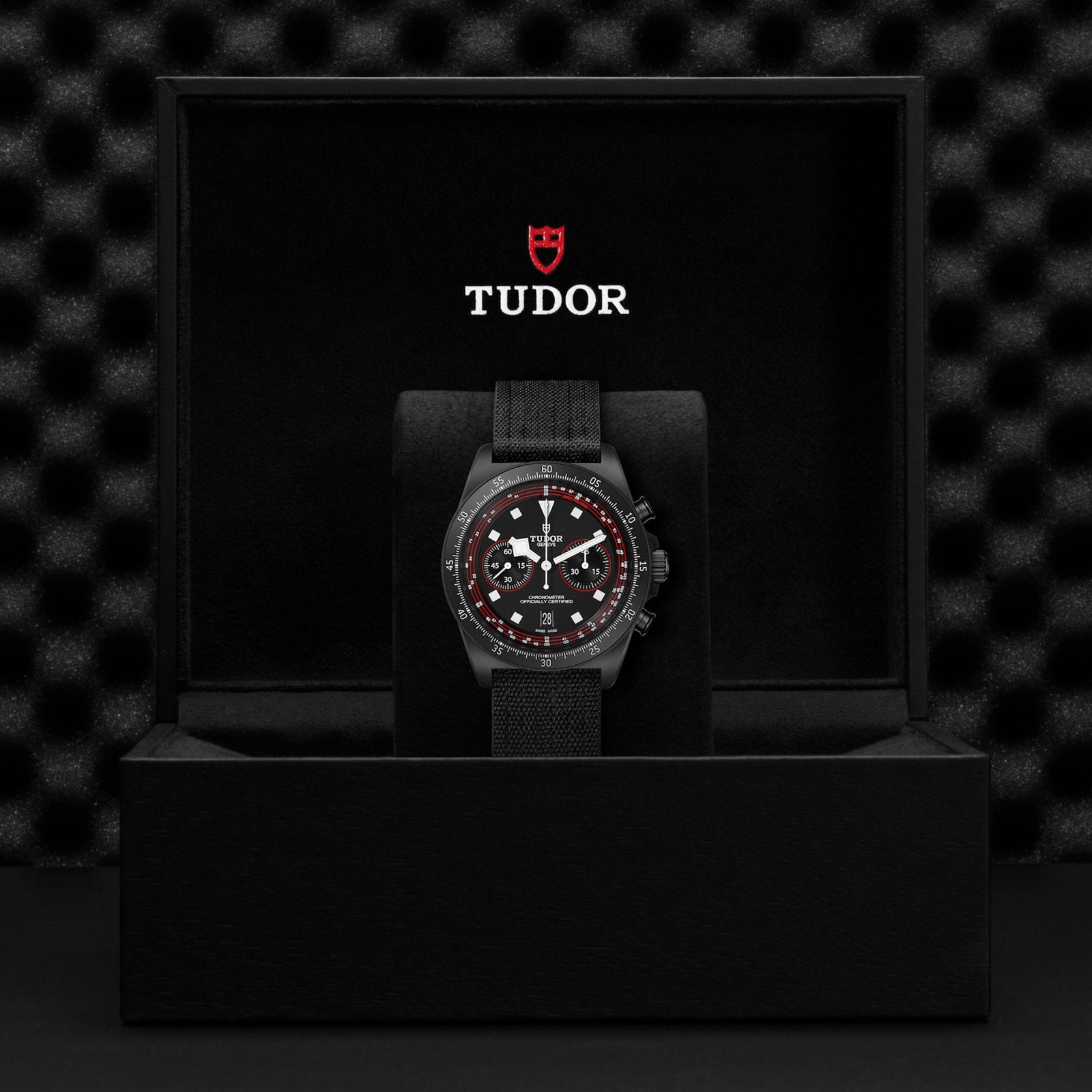 A TUDOR Pelagos FXD Chrono with a black strap and red-accented bezel stands upright in a black TUDOR presentation box, showcasing its cycling-specific tachymeter scale.