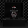 A TUDOR Pelagos FXD Chrono with a black strap and red-accented bezel stands upright in a black TUDOR presentation box, showcasing its cycling-specific tachymeter scale.