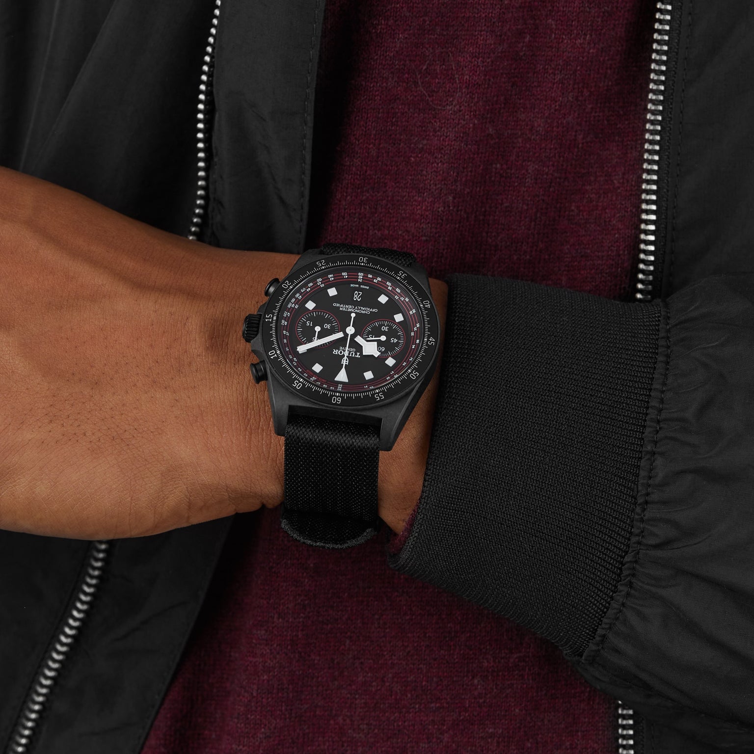 A person wearing a TUDOR Pelagos FXD Chrono wristwatch with a cycling-themed chronograph, dressed in a dark jacket over a maroon top.