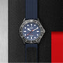 The TUDOR Pelagos FXD dive watch, featuring a blue dial, red seconds hand, and blue fabric strap, is shown on a textured black surface—showcasing its waterproof design ideal for underwater navigation.