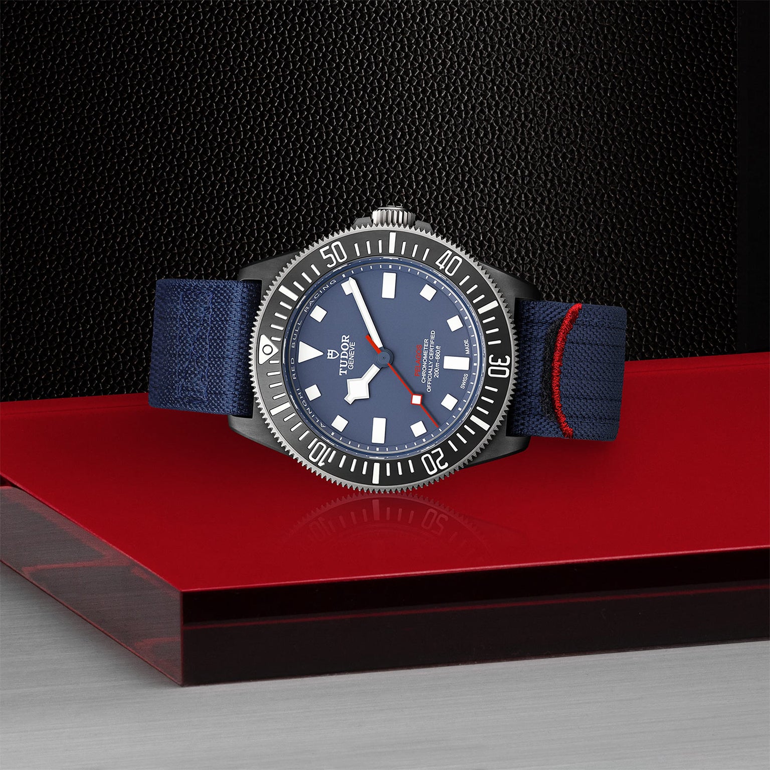 A TUDOR Pelagos FXD wristwatch with a blue face and bezel, white markers, and a blue fabric strap featuring a red stripe, displayed on a red and black surface—ideal for underwater navigation.