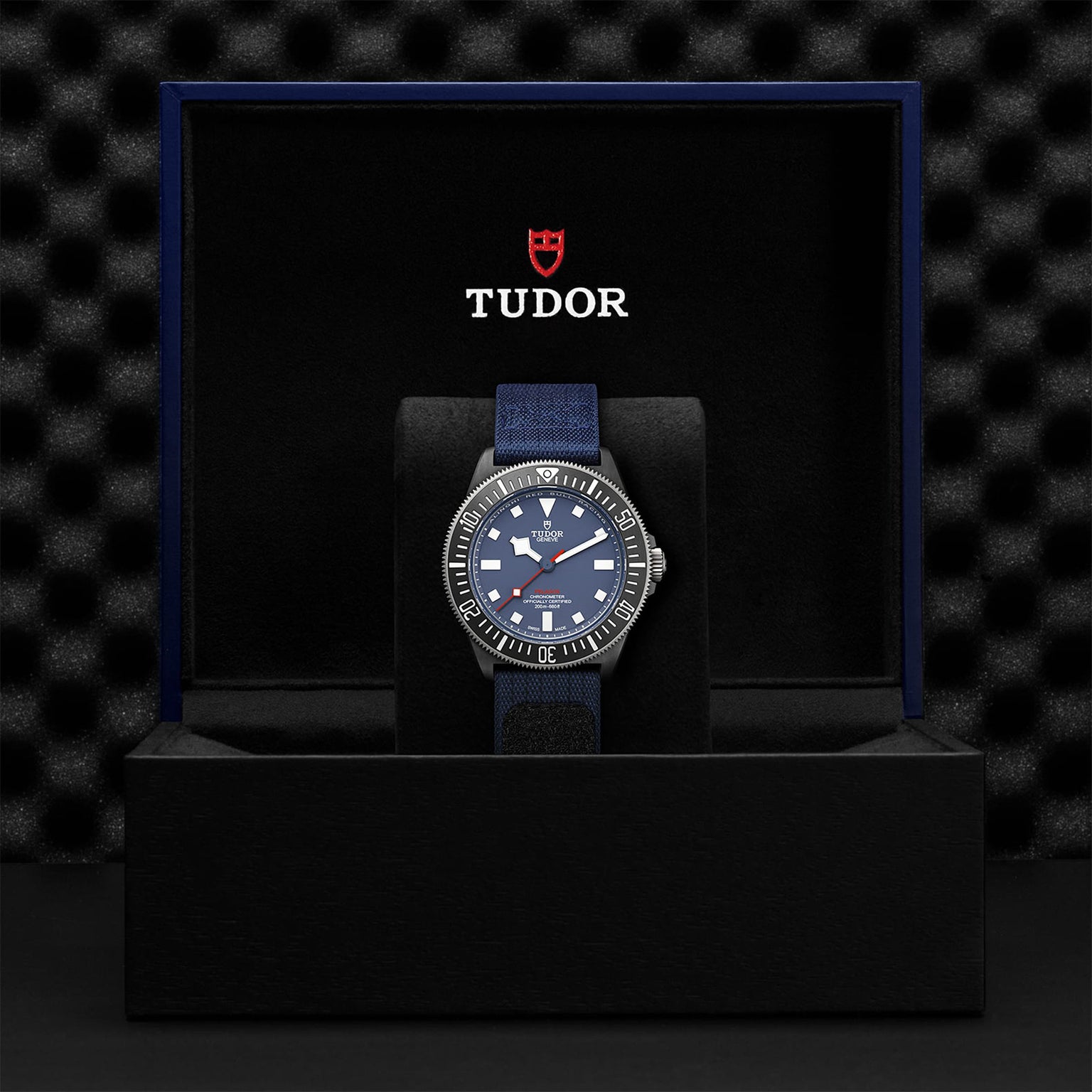 A TUDOR Pelagos FXD with a blue dial and fabric strap is shown in an open black TUDOR box against a textured black background.