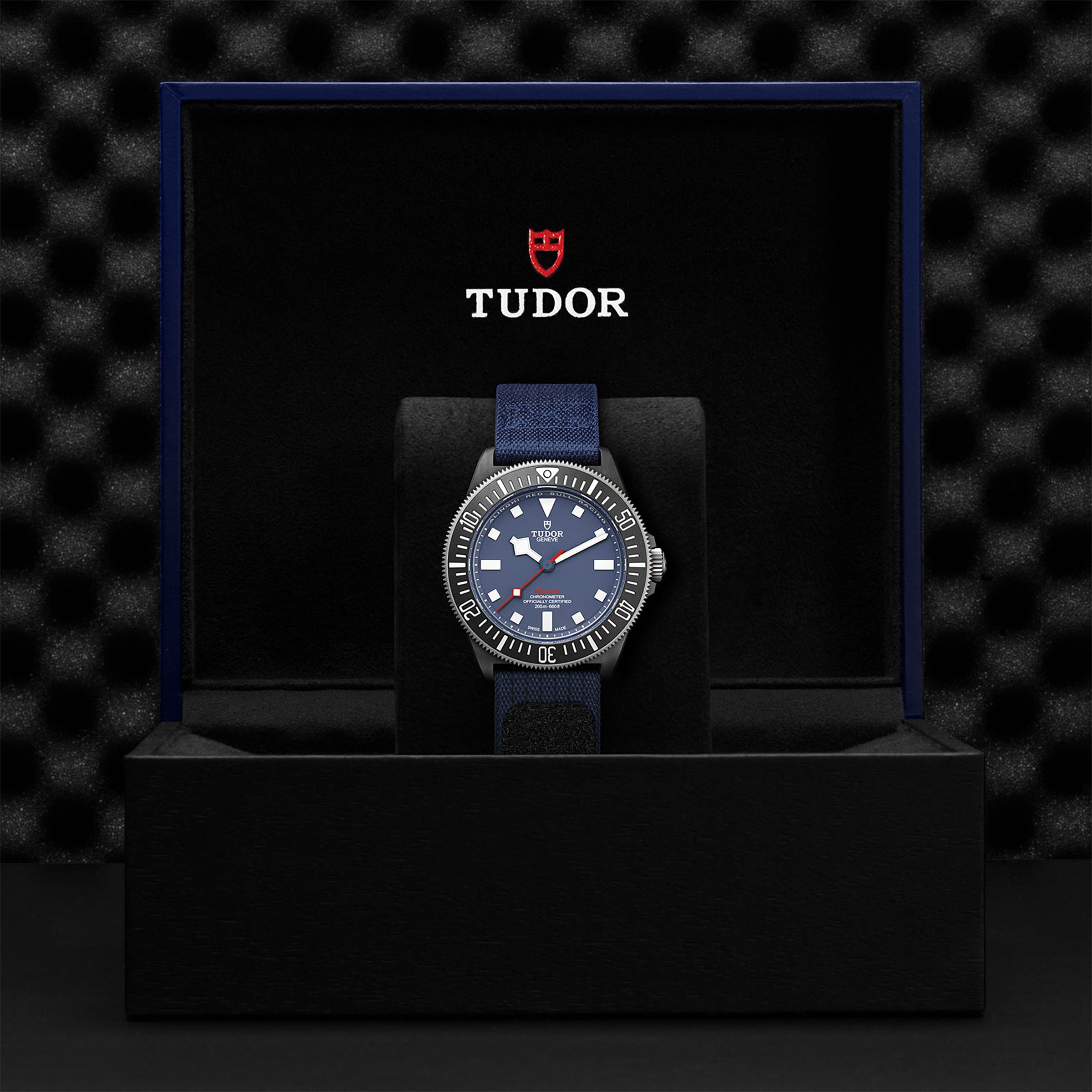 A TUDOR Pelagos FXD with a blue dial and fabric strap is shown in an open black TUDOR box against a textured black background.