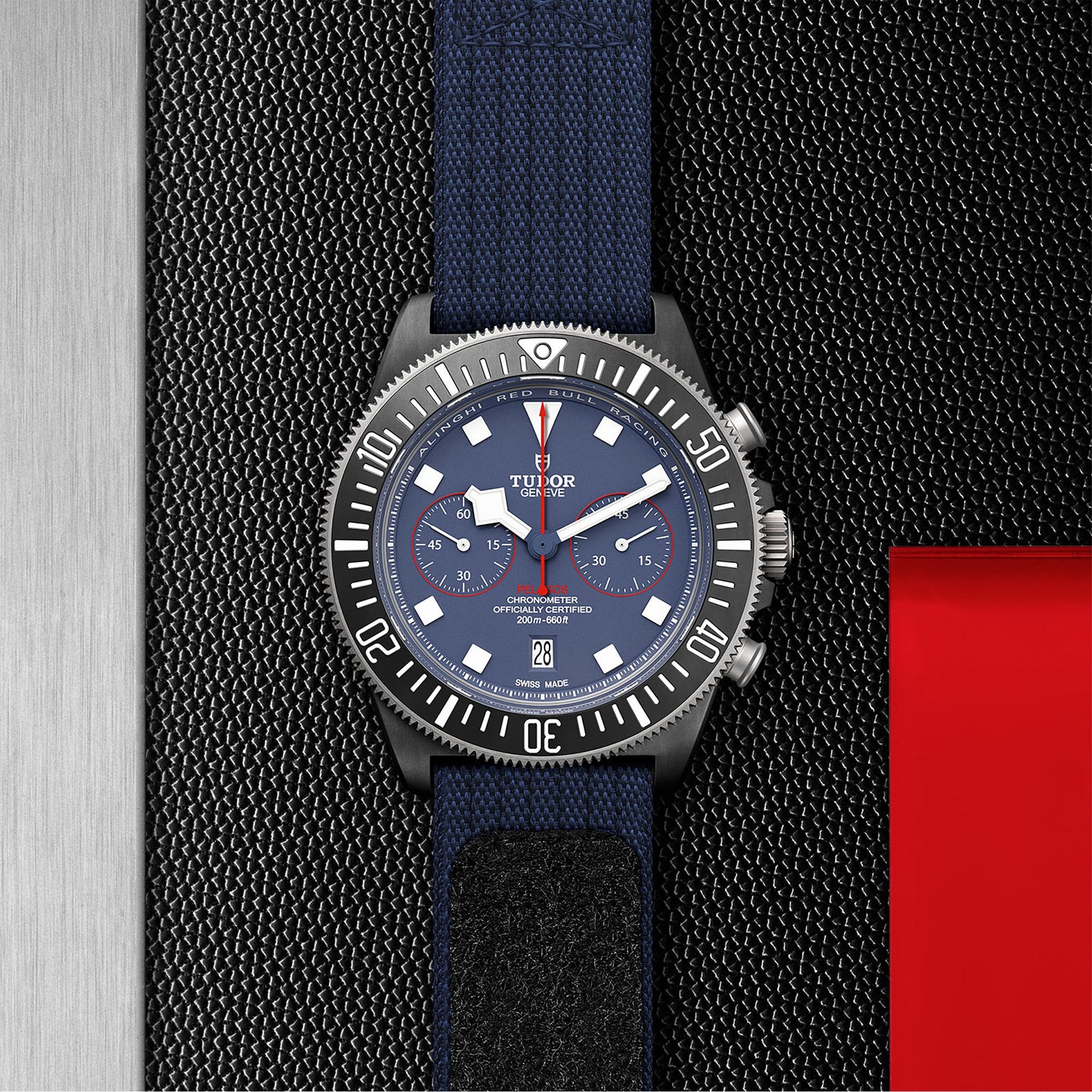 A TUDOR Pelagos FXD Chrono wristwatch, inspired by French Navy navigation, features a blue strap and dial with white and red accents plus three subdials, showcased on a textured black surface next to metallic and red panels.