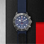 A TUDOR Pelagos FXD Chrono wristwatch, inspired by French Navy navigation, features a blue strap and dial with white and red accents plus three subdials, showcased on a textured black surface next to metallic and red panels.