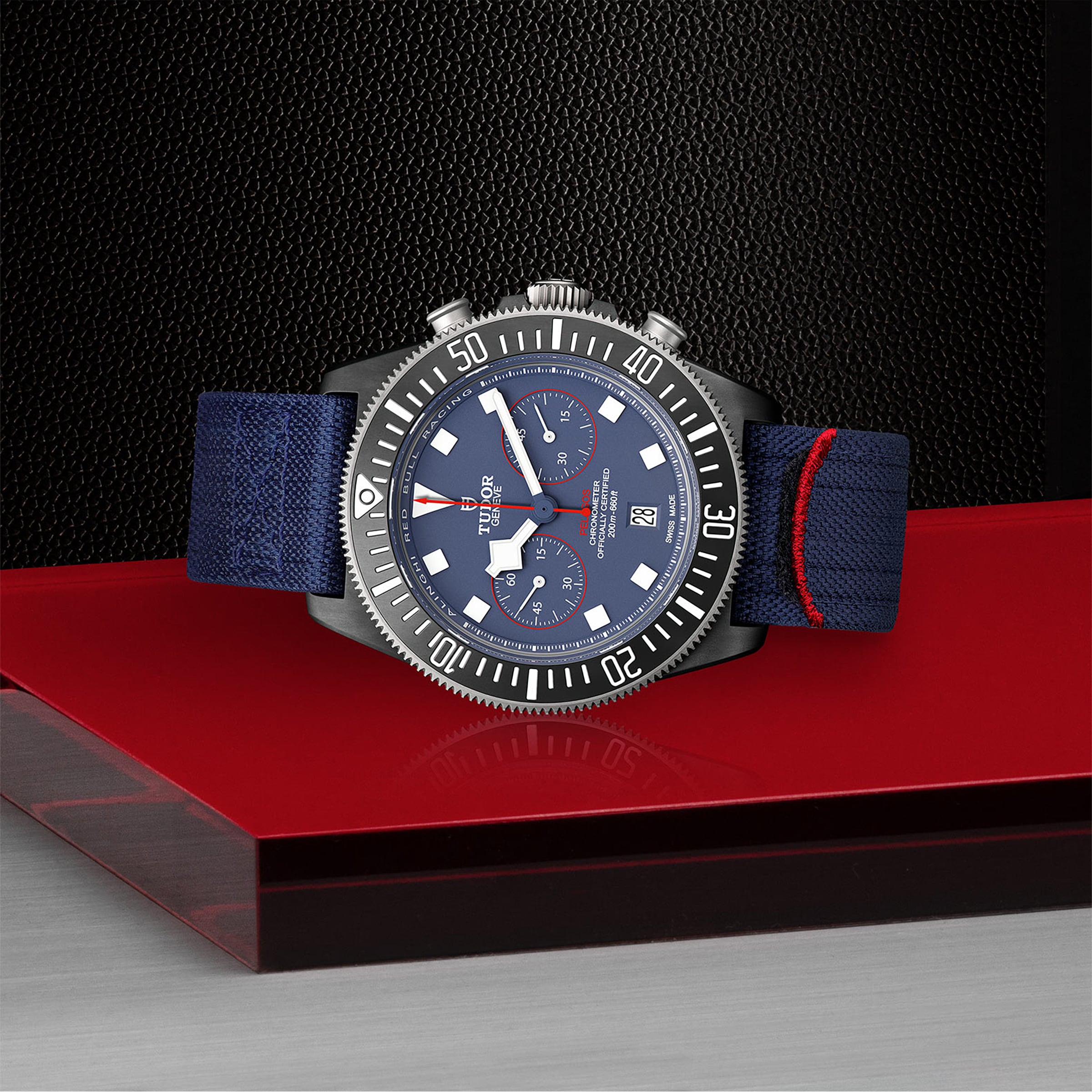 The TUDOR Pelagos FXD Chrono, a blue chronograph inspired by the French Navy, lies on a red acrylic platform with its blue fabric strap highlighted against a textured black background.