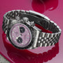 A TUDOR Black Bay Chrono is displayed on a reflective pink surface, featuring a pink dial with black subdials and showcasing its Swiss chronometer-certified precision.