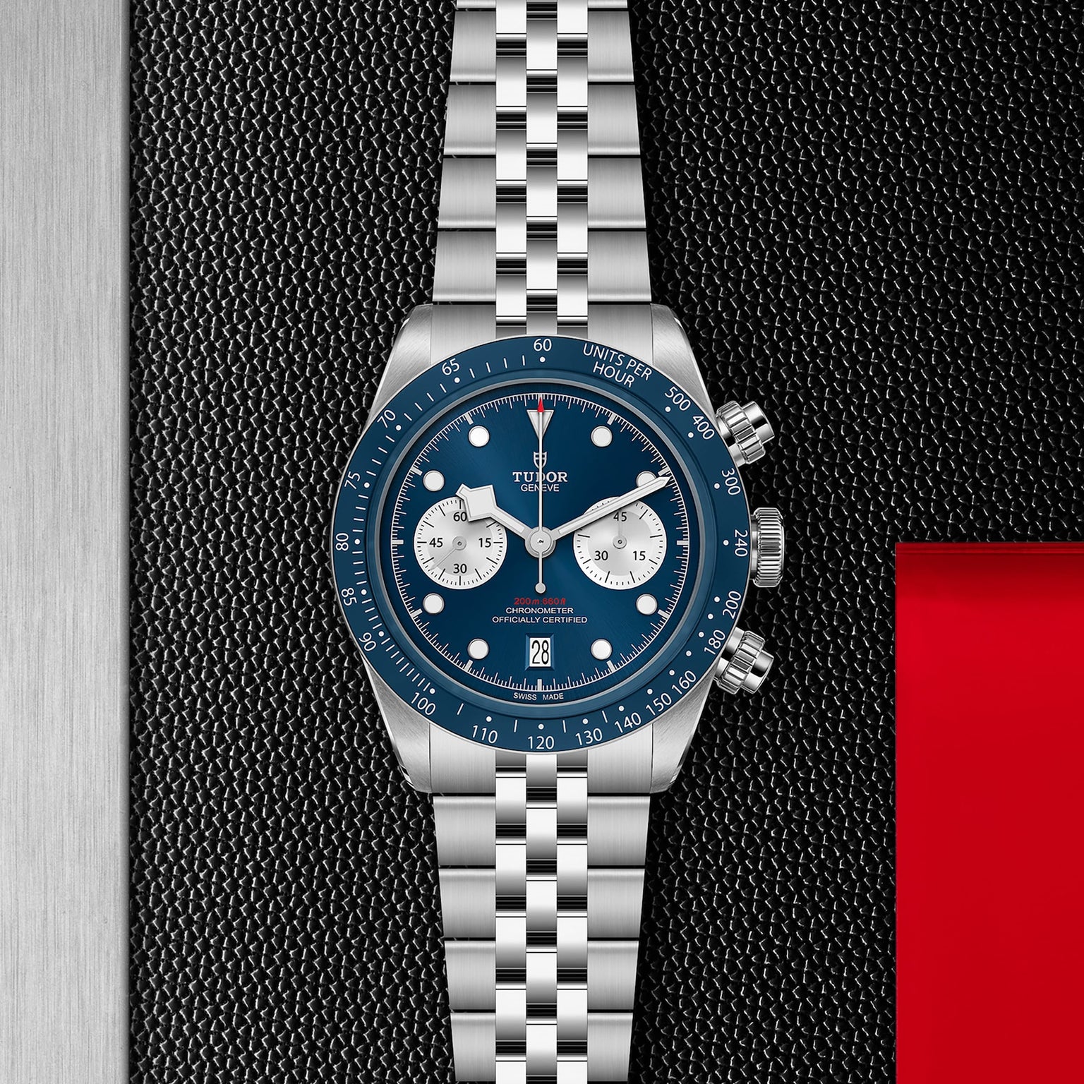 The TUDOR Black Bay Chrono wristwatch, featuring a blue dial and bezel, silver subdials, and a jubilee-style bracelet, is displayed on a textured black and metallic background. Available at Tudor Boutiques.
