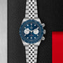 The TUDOR Black Bay Chrono wristwatch, featuring a blue dial and bezel, silver subdials, and a jubilee-style bracelet, is displayed on a textured black and metallic background. Available at Tudor Boutiques.
