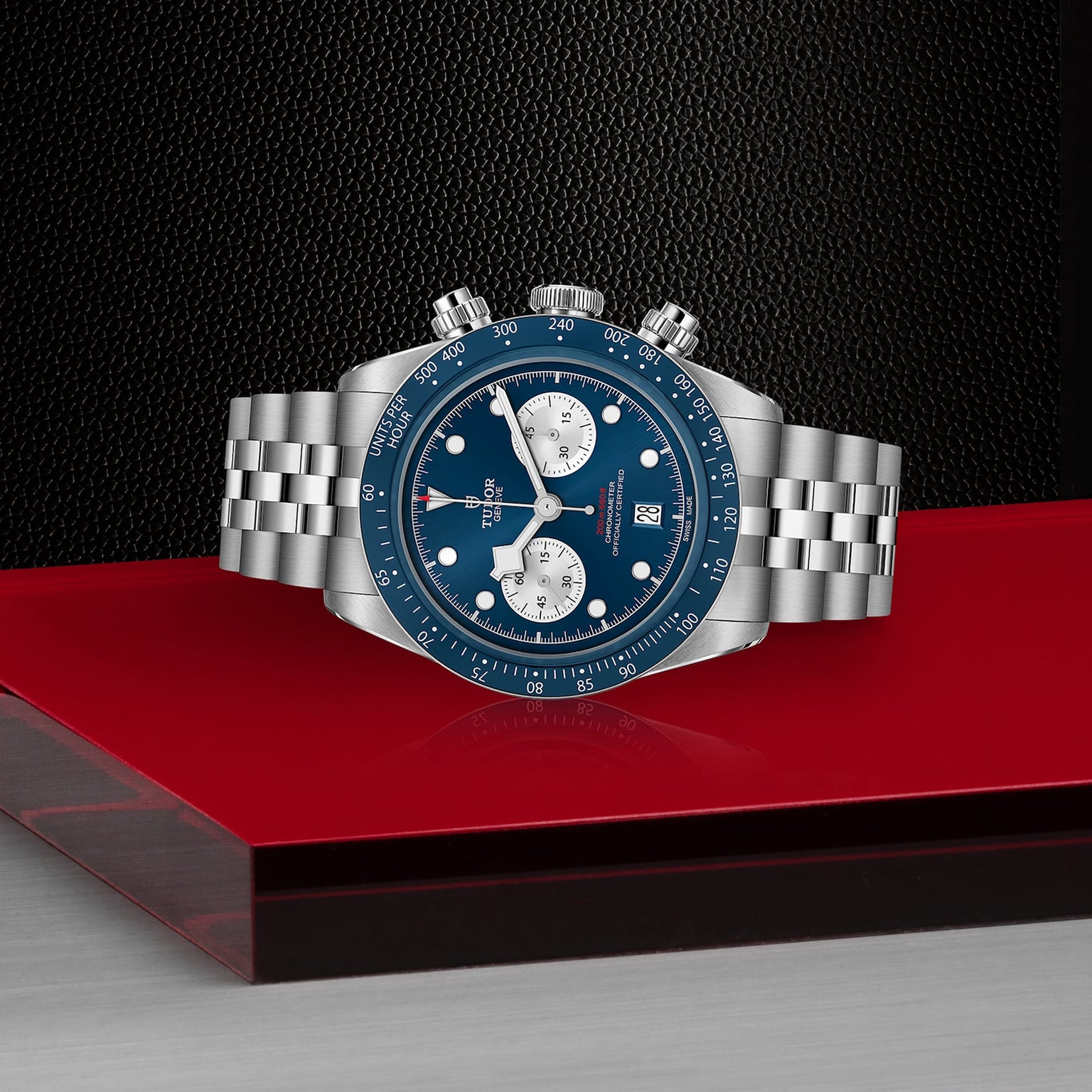 The TUDOR Black Bay Chrono features a stainless steel case, blue bezel and dial with white subdials, date display, and a five-link bracelet—displayed on red and gray—available at select Tudor Boutiques.