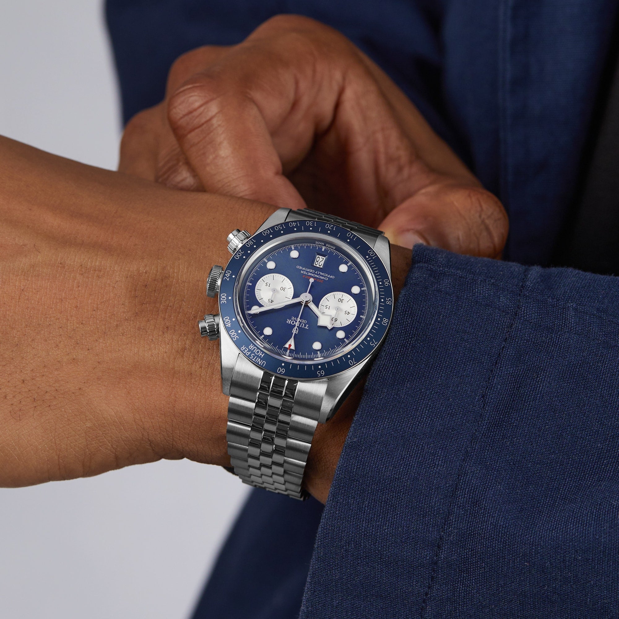 A person in a blue jacket adjusts a TUDOR Black Bay Chrono silver wristwatch with a blue face and white subdials.