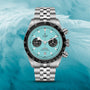 The TUDOR Black Bay Chrono with a turquoise dial, black bezel, and jubilee bracelet is displayed against a light blue feather background, highlighting its Swiss Chronometer precision.