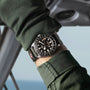 A person in a dark green jacket showcases the TUDOR Pelagos FXD GMT, a black wristwatch with a rotating bezel and fabric strap.