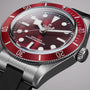 Close-up of the TUDOR Black Bay 58 wristwatch featuring a red bezel, burgundy dial, and black rubber strap on a gray background.