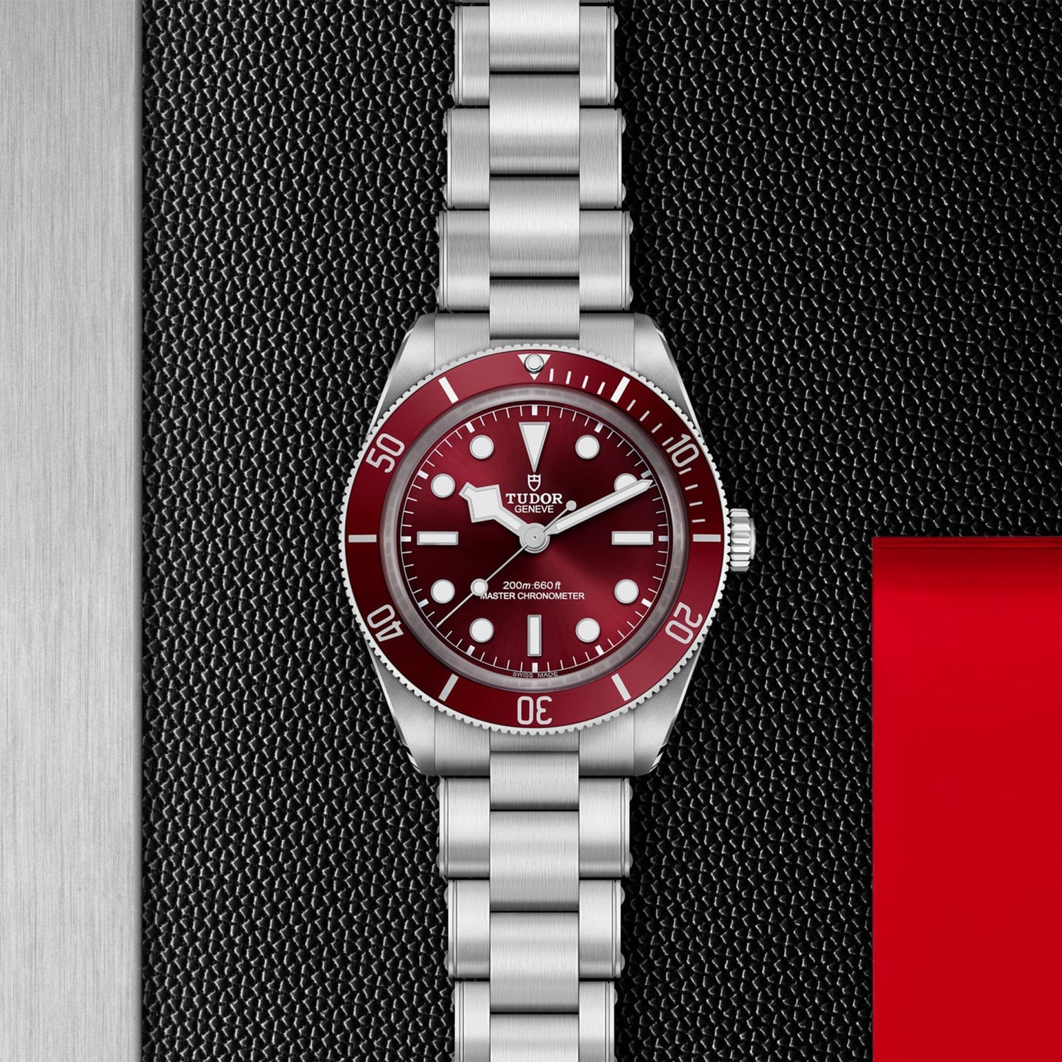 TUDOR Black Bay 58 stainless steel watch with a red bezel and red dial, shown on a textured black background.