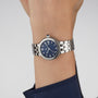 A TUDOR Clair de Rose silver wristwatch with a blue dial and Roman numerals, worn on the wrist of someone in a blue sleeve.