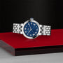 The TUDOR Clair de Rose, a silver watch with a blue face and Roman numerals, is displayed on a red and black surface.