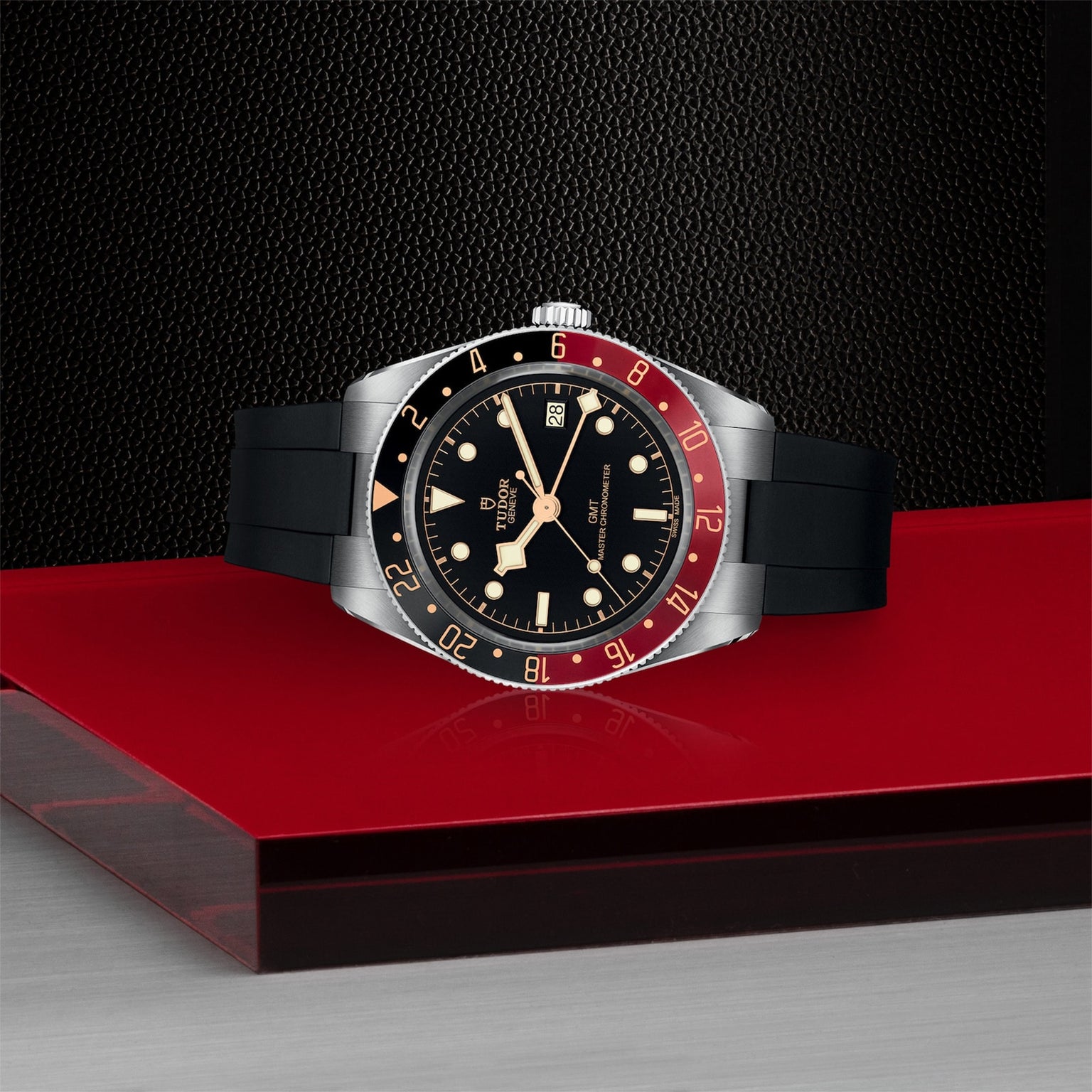 The TUDOR Black Bay 58 GMT, featuring a black dial, red and black bezel, and black rubber strap, is displayed on a red and metallic surface against a textured dark background.