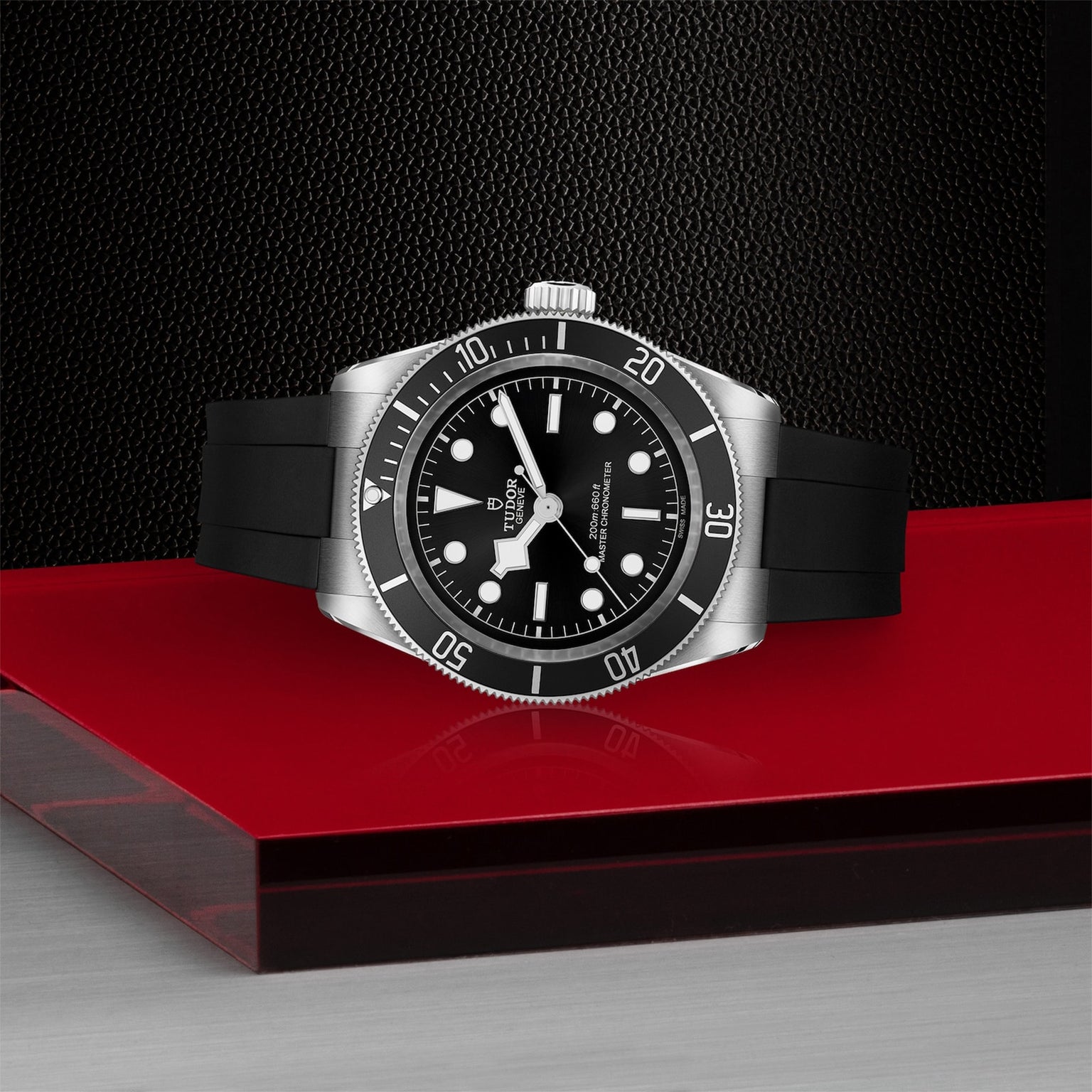 The TUDOR Black Bay is a METAS-certified stainless steel diving watch with a black dial, black bezel, black rubber strap, and white hour markers, shown on a red and silver surface against a dark textured background.
