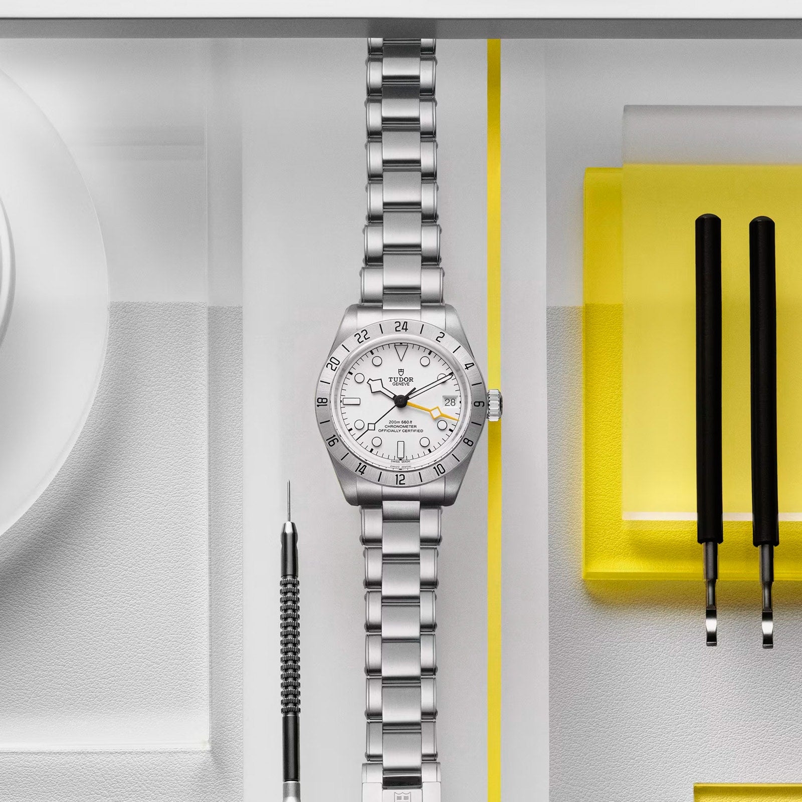 The TUDOR Black Bay Pro wristwatch is displayed beside watchmaking tools on a white and yellow background.