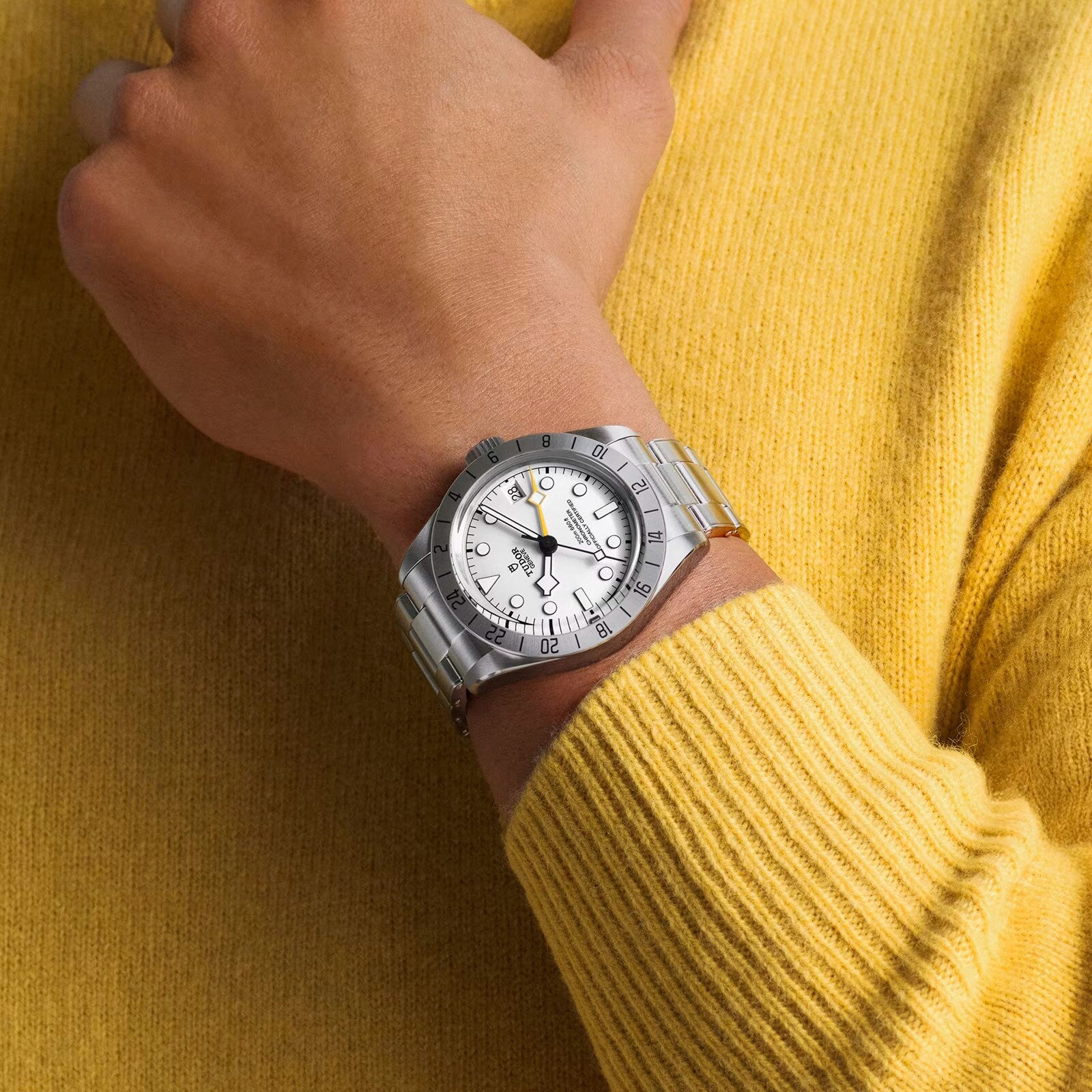 A person wearing a TUDOR Black Bay Pro wristwatch and a yellow sweater, with their hand resting near their wrist.