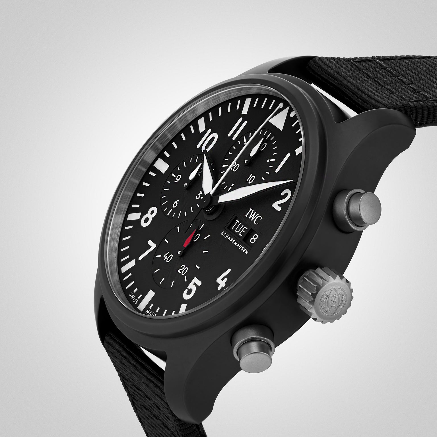 The IWC Schaffhausen Pilots Watch Chronograph Top Gun features a black dial with white numerals, two push buttons, a textured crown, and a black fabric strap, presented on a light gradient background.