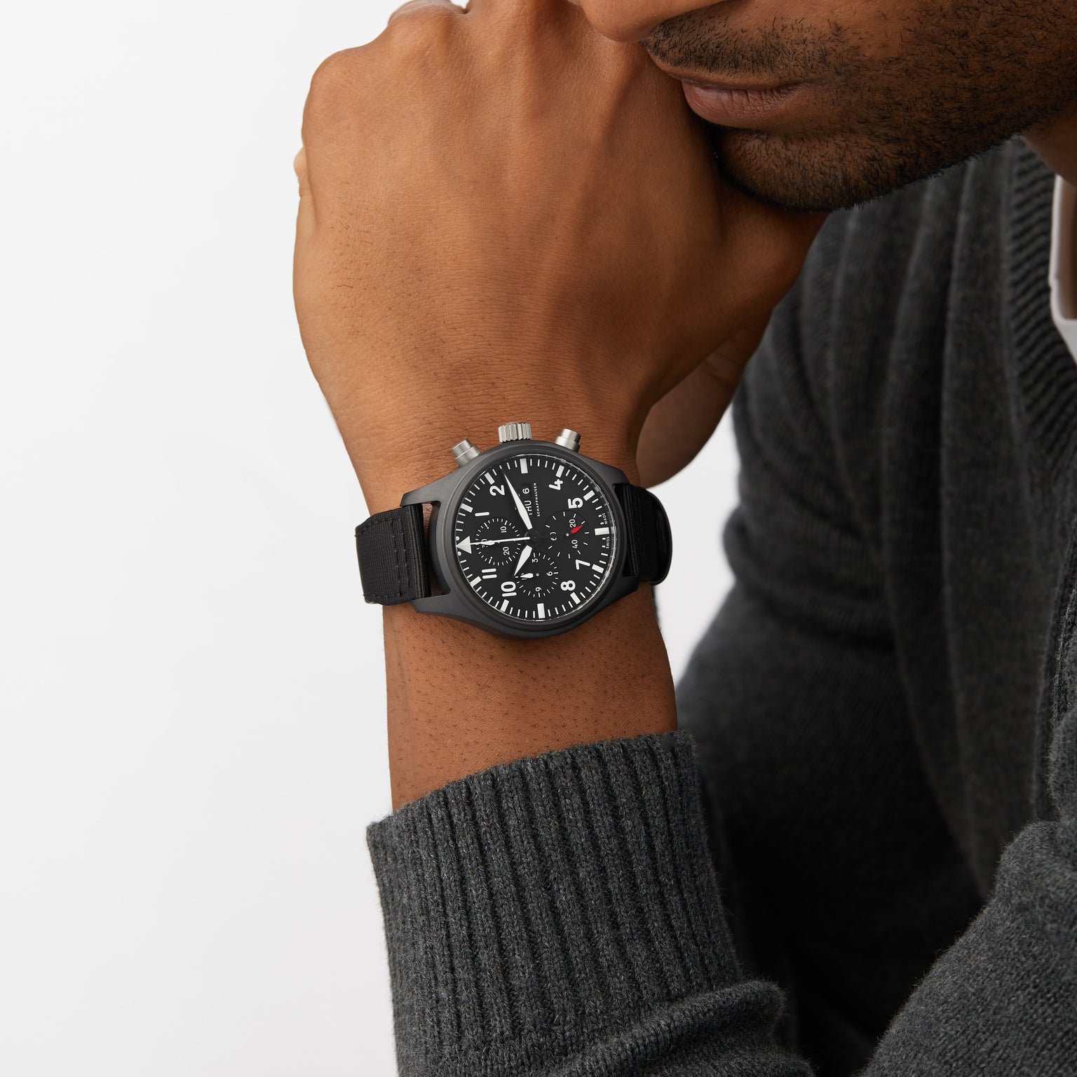 A person in a dark sweater rests their chin on their hand, showcasing the IWC Schaffhausen Pilots Watch Chronograph Top Gun—a black chronograph wristwatch with multiple dials and a black strap, inspired by classic pilot’s watches.