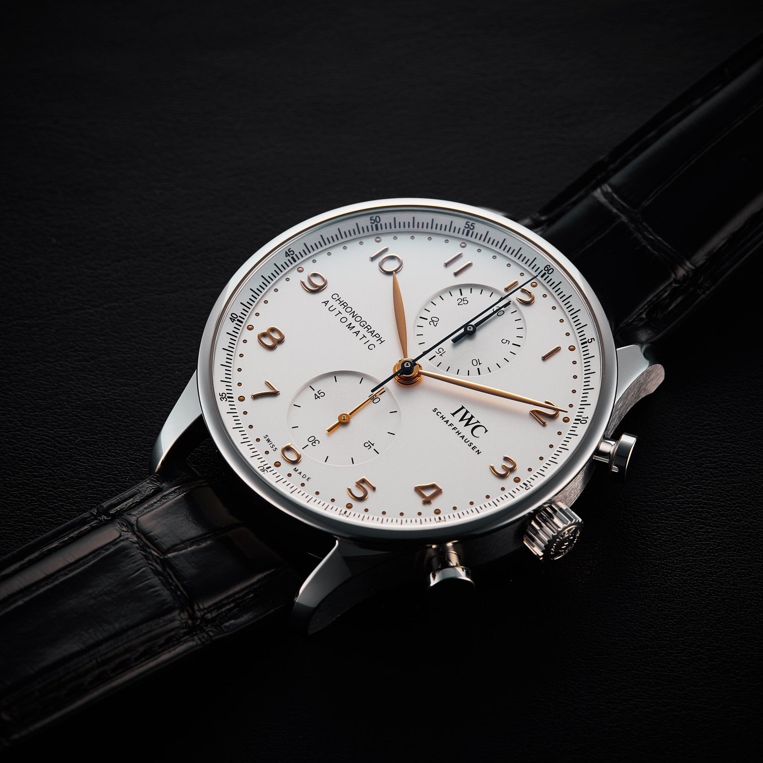 The IWC Schaffhausen Portugieser Chronograph, featuring a silver stainless steel case, white dial with gold numbers, two subdials, and a black leather strap, rests on a black surface.