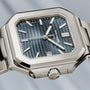 A Patek Philippe Cubitus 7128/1G-001 wristwatch features a silver rectangular case, blue striped dial, and metal bracelet.