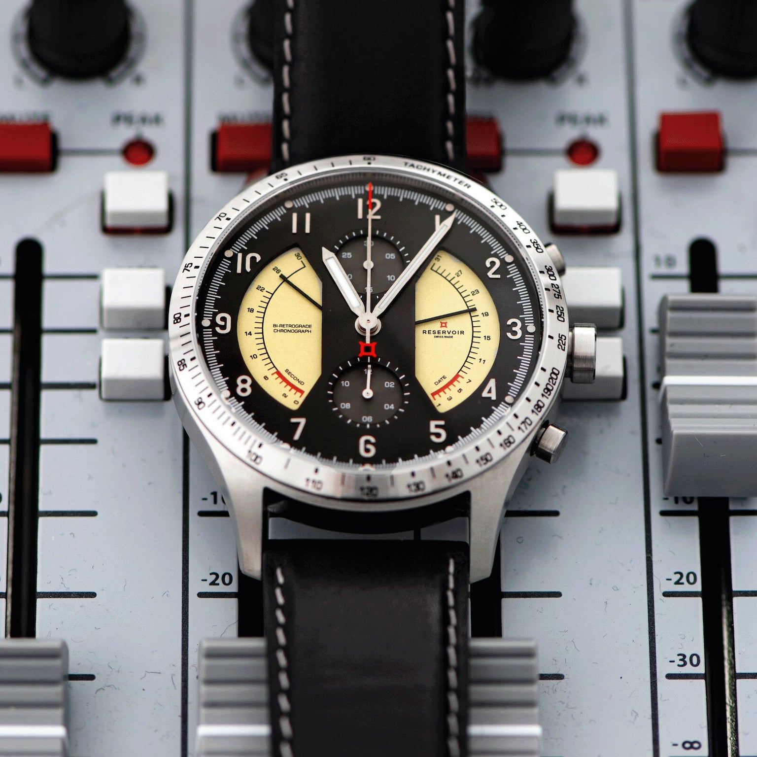 The Reservoir Sonomaster Chronograph Vintage wristwatch with a black leather strap rests on an audio mixing console with sliders and knobs.