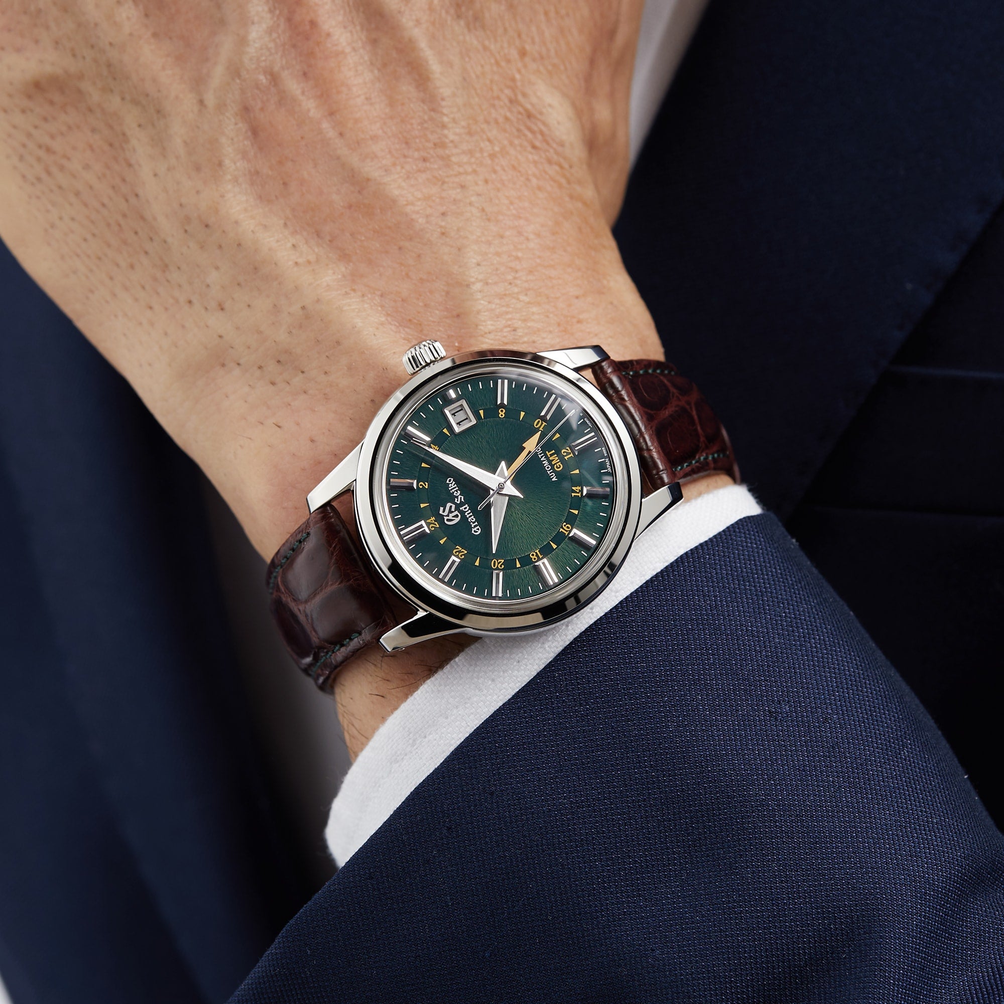 A person in a dark blue suit showcases the Grand Seiko Automatic GMT Elegance, featuring a Mount Iwate dial in British Racing Green, a silver case, and a brown leather strap.
