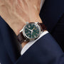 A person in a dark blue suit showcases the Grand Seiko Automatic GMT Elegance, featuring a Mount Iwate dial in British Racing Green, a silver case, and a brown leather strap.
