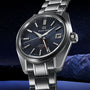 The Grand Seiko Hi-Beat GMT Heritage, a stainless steel wristwatch with a blue textured dial, date display, and GMT hand featuring Zaratsu polishing, is shown against a snowy mountain landscape at night.