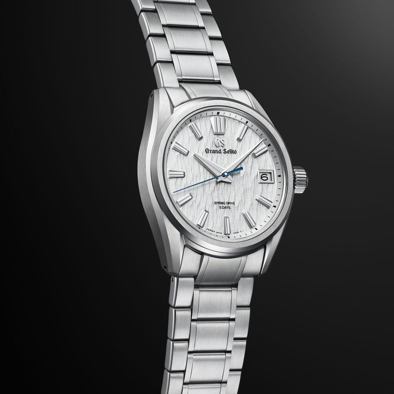 The Grand Seiko Spring Drive 5 Days Evolution 9 is a stainless steel wristwatch with a textured white dial, silver hour markers, date window at 3 o’clock, and Spring Drive movement, set against a dark gradient backdrop.