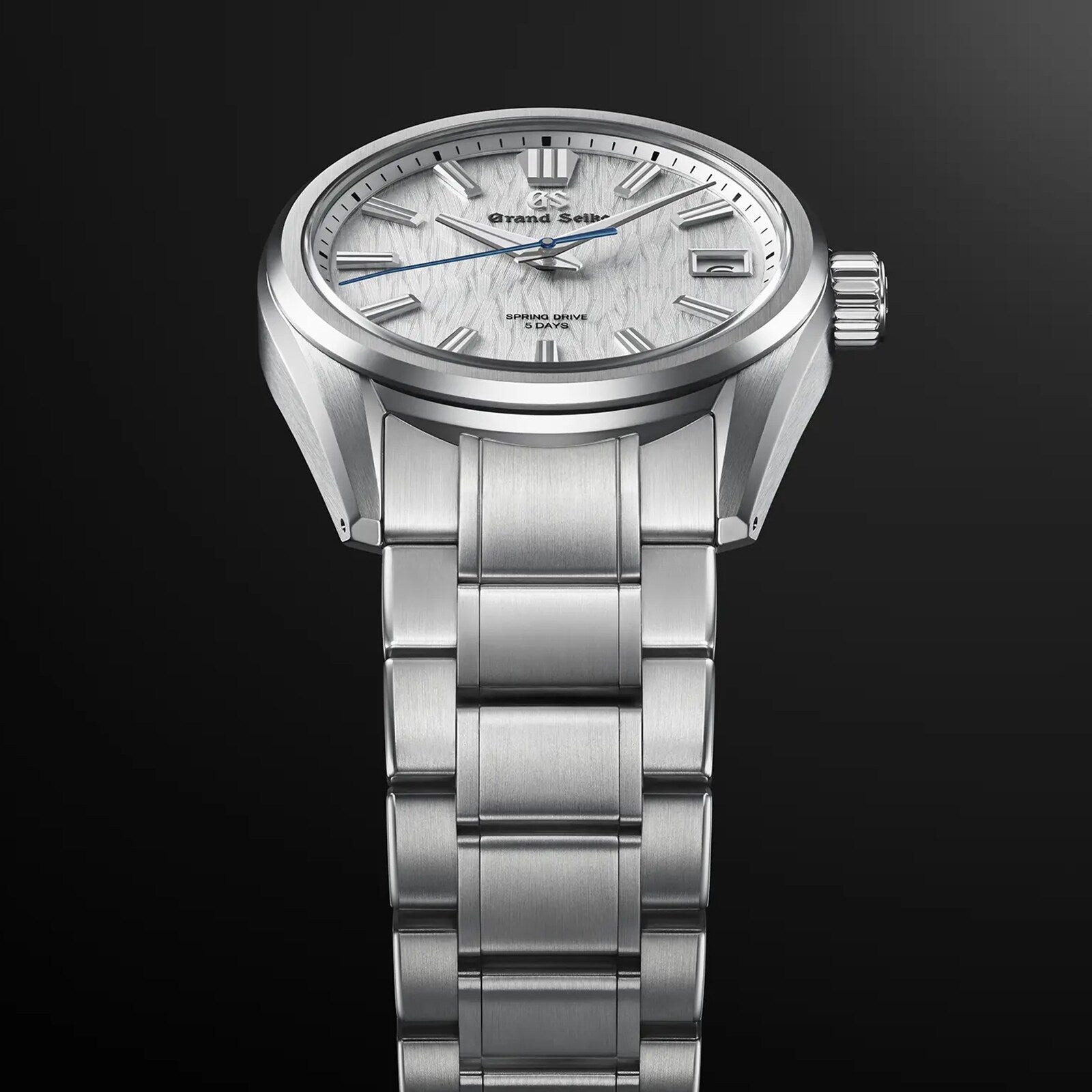 The Grand Seiko Spring Drive 5 Days Evolution 9, featuring a textured white dial and stainless steel bracelet, is elegantly displayed against a dark gradient background.