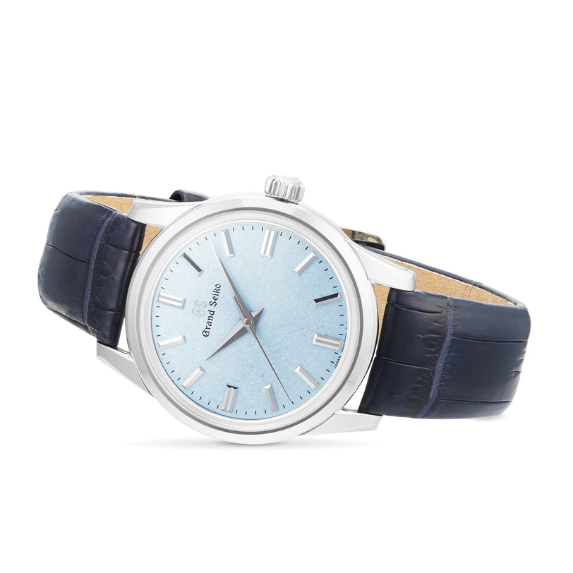 The Grand Seiko Manual Elegance features a silver case, light blue textured dial, silver hour markers, and a dark blue leather strap with the Caliber 9S64 movement, shown at an angle.