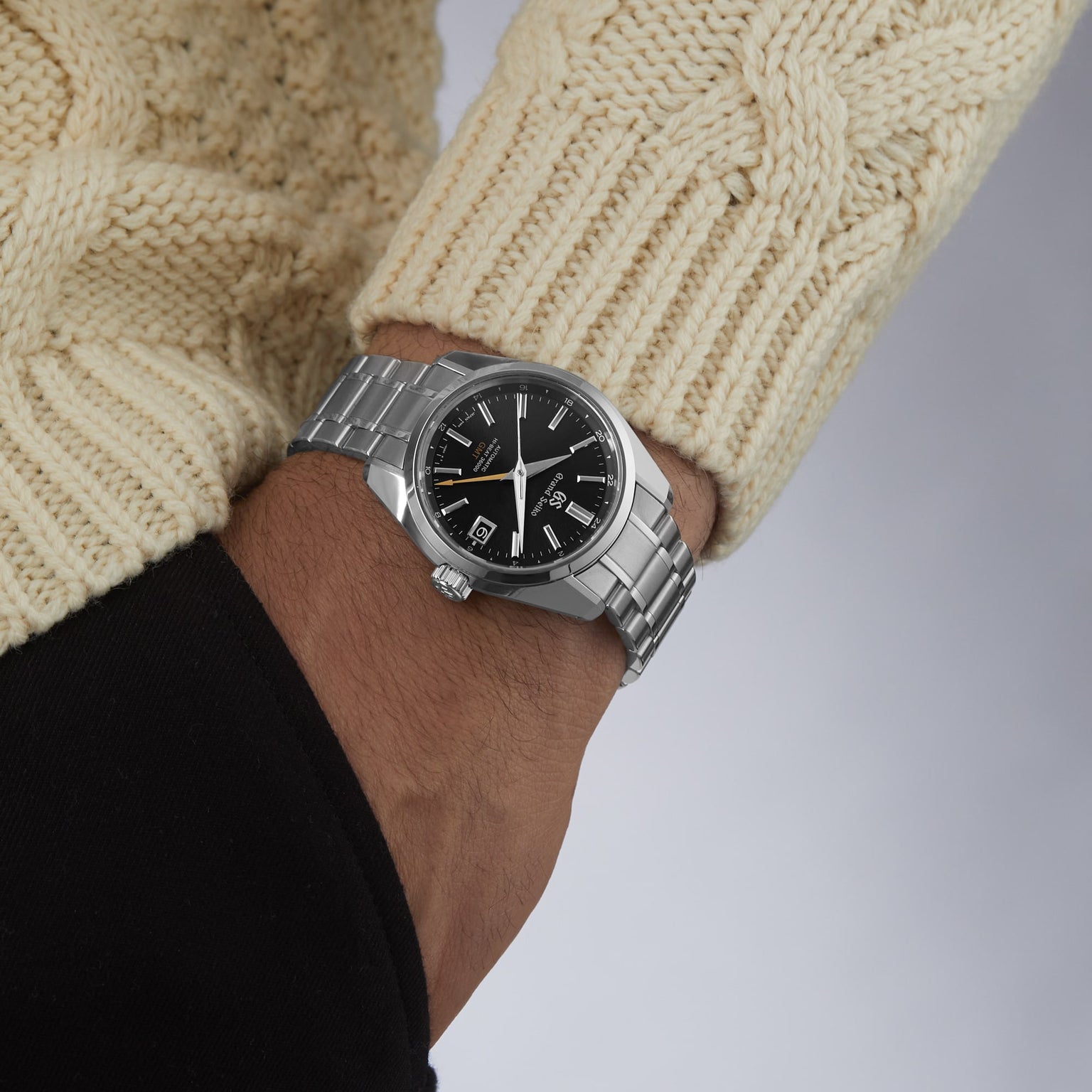 A person in a cream knit sweater and black top is wearing the Grand Seiko Hi-Beat GMT Heritage watch featuring a silver case, black dial, metal band, and precise automatic movement.