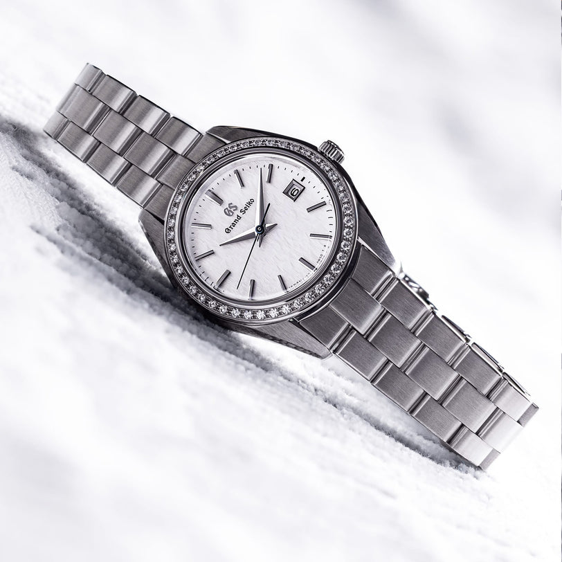 The Grand Seiko Ladies Heritage wristwatch features a stainless steel band, snowflake dial, date window, and 44 diamonds on the bezel, shown on a white surface.
